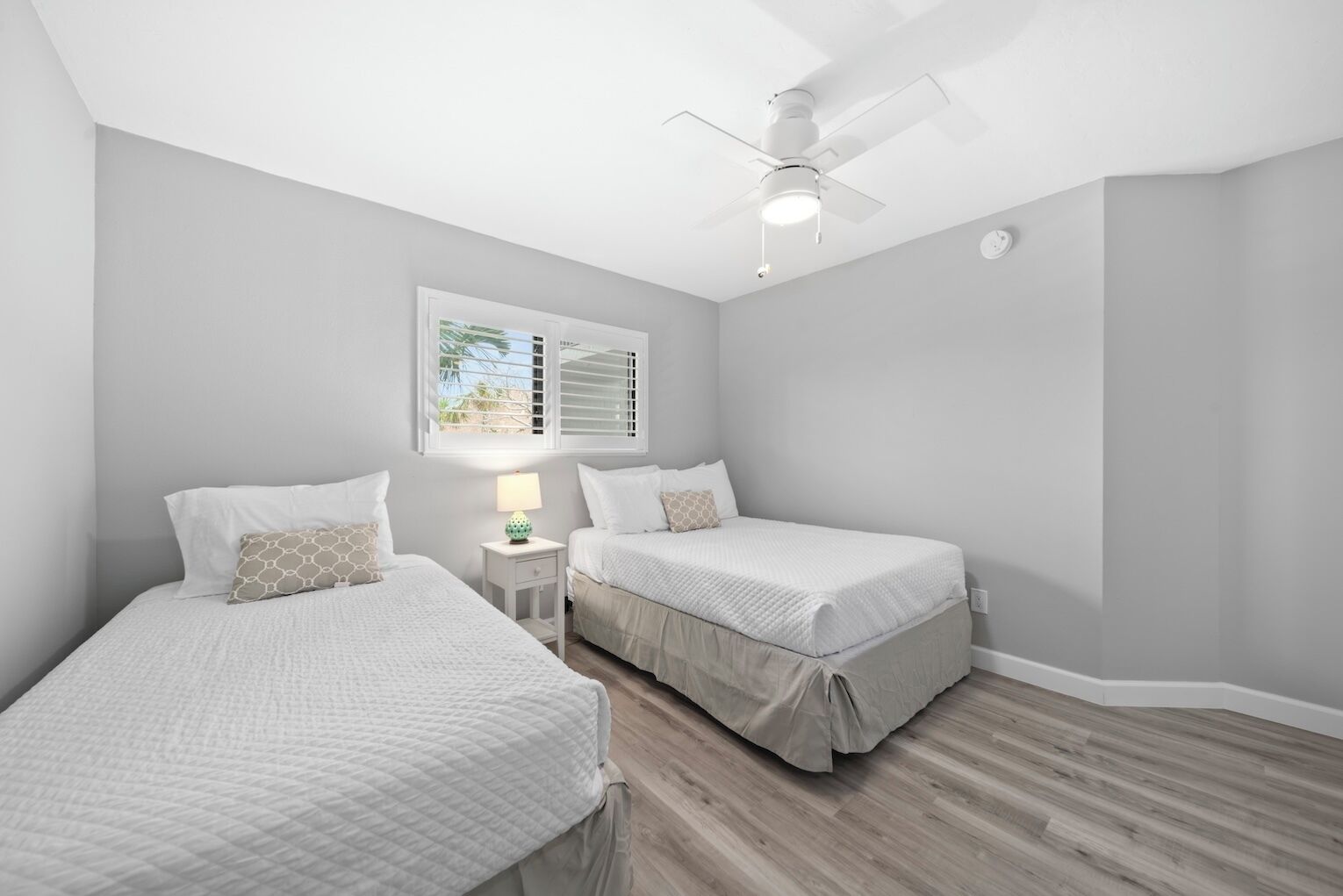 A bedroom with two beds, a ceiling fan, a window with blinds, and a nightstand with a lamp.
