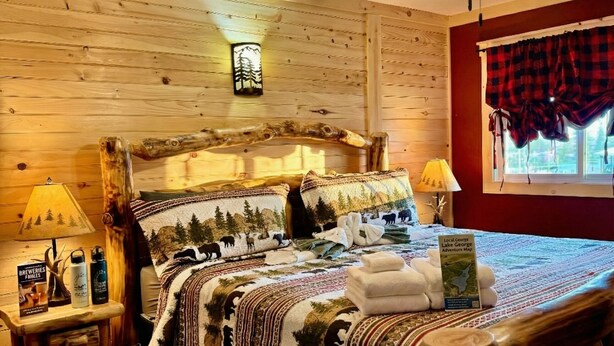 A wooden-paneled room with a bed, a ceiling fan, and a window with curtains. - The Lake George Inn (Lake George)