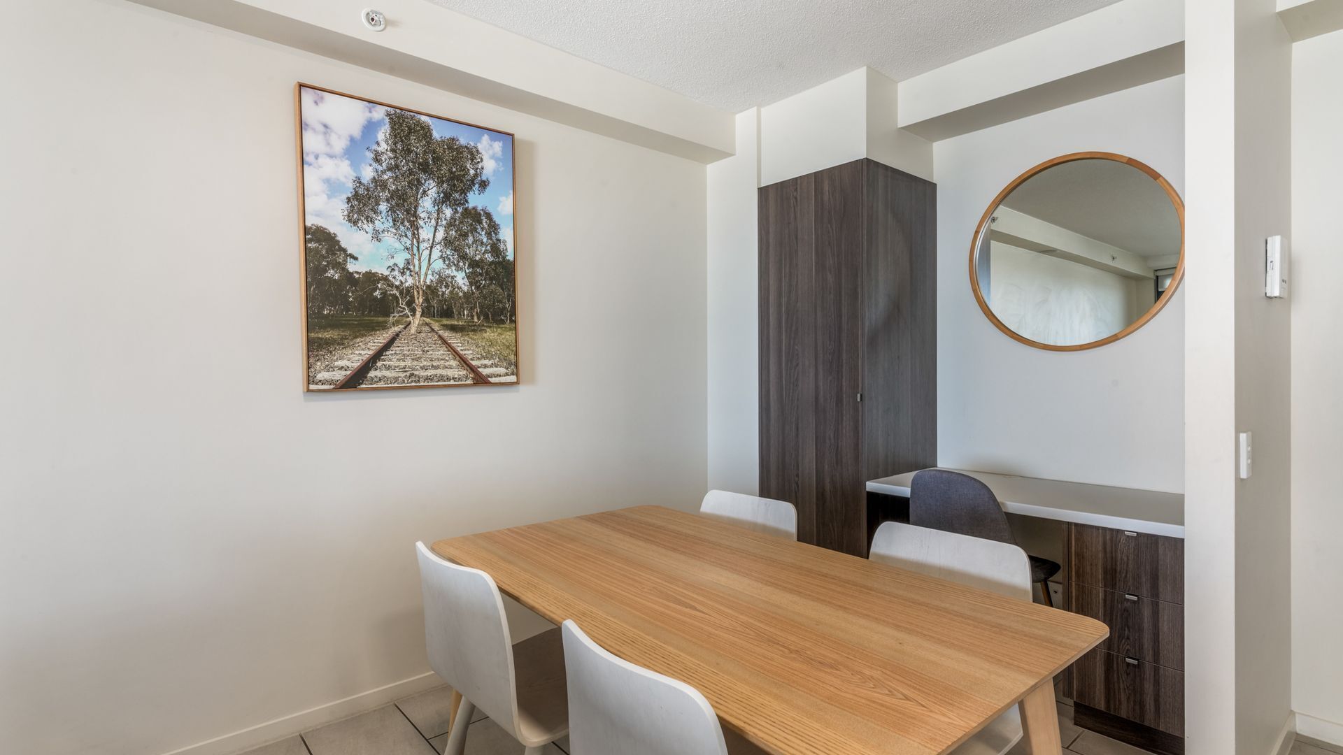 A dining area with a wooden table and white chairs, a dark wood cabinet, a round mirror, and a framed picture of a tree on a railway track.