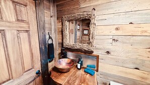 Een rustieke houten badkamer met een houten deur, een spiegel met een geweven lijst, een houten wasbak en een zeepfles.