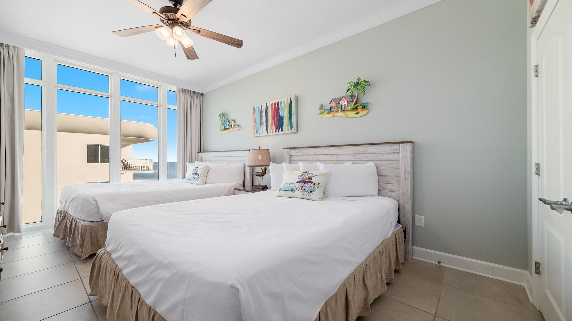 A hotel room with two beds, a ceiling fan, a balcony with a view, and artwork on the walls.
