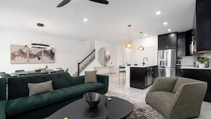 Una sala de estar moderna con un sofá verde, una mesa redonda y un sillón gris. Se observa una zona de cocina con armarios negros y electrodomésticos de acero inoxidable.