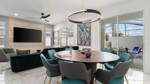 Una sala moderna con una mesa de comedor redonda, sillas color turquesa, un sofá verde, un televisor de pantalla plana y un ventilador de techo.