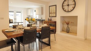 Een eetruimte met een houten tafel gedekt voor vier personen, omringd door stoelen, een grote wandklok en een vaas met gedroogde bloemen.