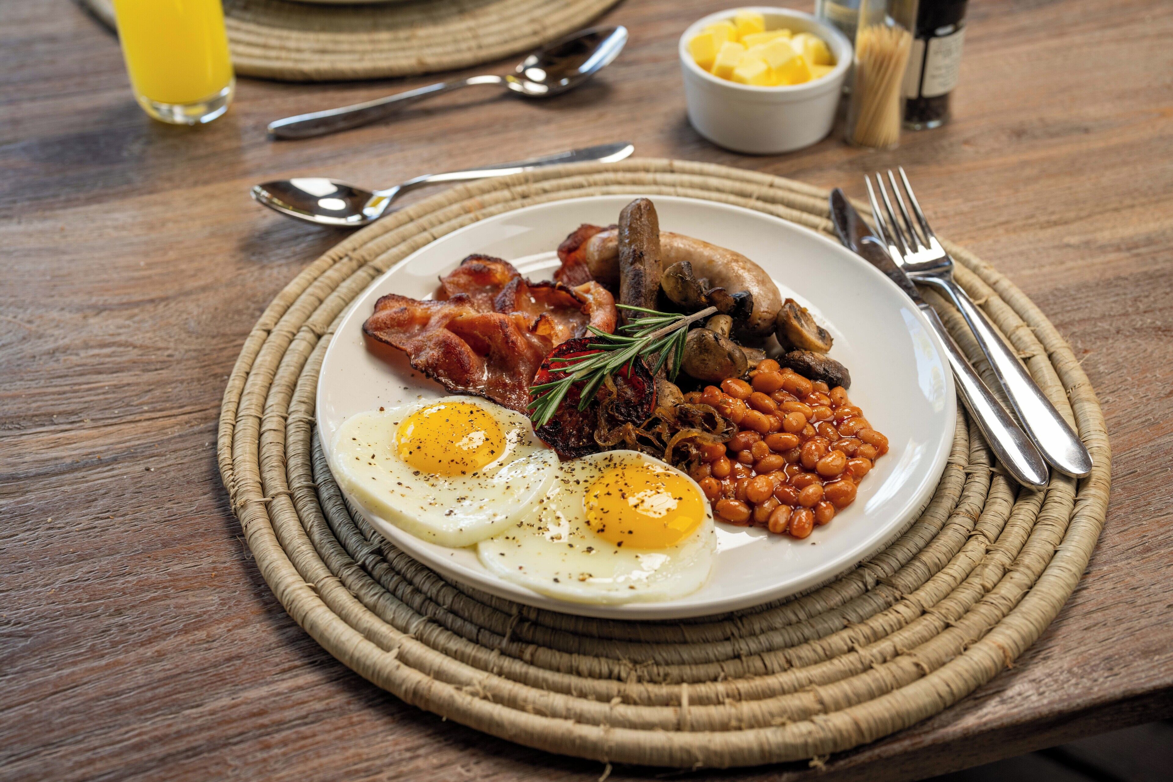 A plate of breakfast with eggs, bacon, mushrooms, beans, and a sprig of rosemary.