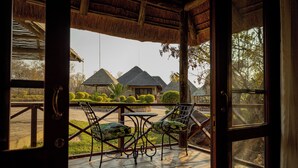 An open door leading to a patio with a table and two chairs, overlooking a thatched-roof building and neatly trimmed bushes. - Room in Guest Room - Mmakuba Private Game Lodge, Deluxe Chalet 7C (Mookgopong)