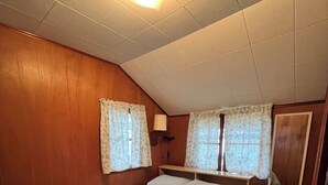 Un dormitorio con cama individual, paredes de madera, una lámpara en el techo, dos ventanas con cortinas, una mesita de noche con lámpara y un espejo en la pared.