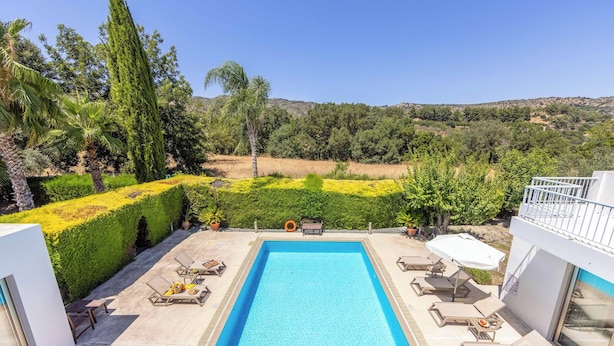 A rectangular swimming pool with lounge chairs and umbrellas, surrounded by well-maintained hedges and trees, with a view of a hilly landscape. - Villa Zoey - Three Bedroom Villa, Sleeps 6 (Theletra)