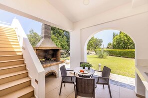 Une terrasse extérieure dotée d’une cheminée en briques, d’une table à manger avec des chaises et offrant une vue sur un jardin bien entretenu.