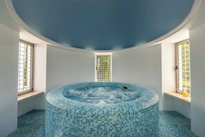 Un jacuzzi circulaire doté d’un sol et de murs recouverts de mosaïques, entouré de fenêtres à cadres blancs et surmonté d’un plafond bleu.