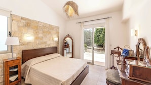 A bedroom with a stone wall, a bed, a nightstand, a dresser, a mirror, and a view of the outdoors. - Villa Zoey - Three Bedroom Villa, Sleeps 6 (Theletra)