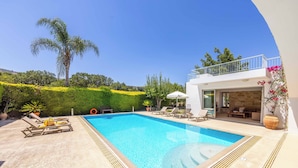 Ein rechteckiger Swimmingpool mit glasklar blauem Wasser, umgeben von einer Betonplattform. Liegestühle und ein Tisch mit einem gelben Handtuch darauf sind vorhanden. Ein weißes Gebäude mit Balkon und Steinmauer grenzt an den Pool.