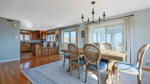 Un comedor amplio con una gran mesa de madera y sillas, una araña y vistas al exterior a través de ventanas grandes.