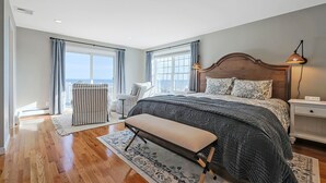 Un dormitorio con una cama grande, un banco, una mesita de noche y una lámpara. Hay dos ventanas con cortinas, vistas al exterior y un respiradero en el techo.