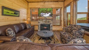 A cozy log cabin interior with a stone fireplace, leather sofas, and a large window offering a view of the outdoors. - *Gorgeous 3BD Cabin w/ VIEWS! Hot Tub + Games!* (Sevierville)