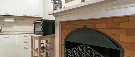 A kitchen with white cabinets, a microwave, a wooden shelf with bottles, and a fireplace with decorative ironwork.
