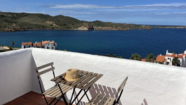 Une terrasse avec une table en bois et un chapeau de paille, d’où l’on peut admirer un paysage côtier avec des collines et une étendue d’eau.
