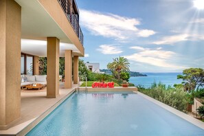 A modern pool area with a clear view of the sea, surrounded by greenery and a white building with a balcony.