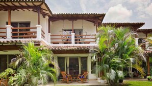 A two-story house with a balcony, a patio with chairs, and lush greenery.