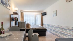 A hotel room with a bed, a desk with a chair, a bookshelf, a TV mounted on the wall, and a window with curtains. - Ferienwohnung in St. Peter-Ording (St. Peter-Ording)