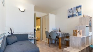 A cozy living area with a grey sofa, wooden dining table, and chairs. A wooden bookshelf is against the wall, and a kitchen is visible in the background. - Ferienwohnung in St. Peter-Ording (St. Peter-Ording)