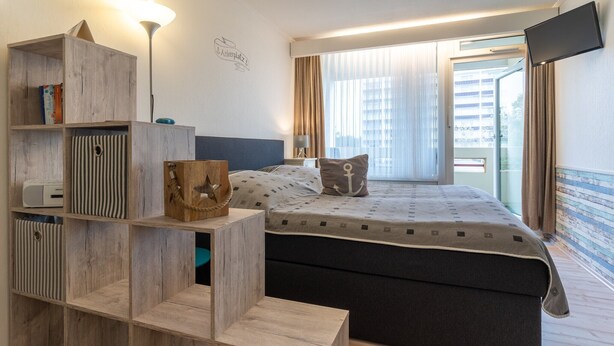 A hotel room with a bed, a TV mounted on the wall, a wooden bookshelf, and a window with curtains. - Ferienwohnung in St. Peter-Ording (St. Peter-Ording)