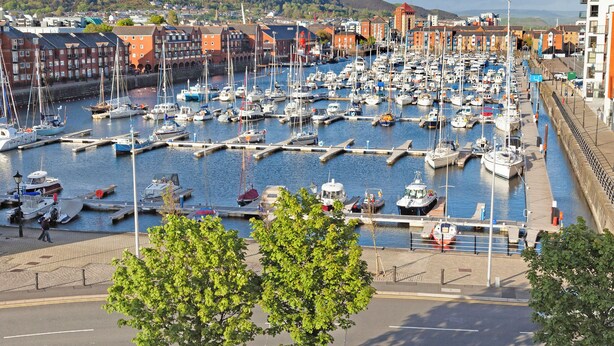 Un port de plaisance où sont amarrés de nombreux bateaux, entouré de bâtiments et avec un paysage vallonné en arrière-plan.