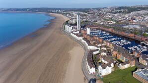 Une vue aérienne d’une ville côtière, avec une longue plage de sable, une marina où sont amarrés de nombreux bateaux, et un mélange de bâtiments résidentiels et commerciaux.