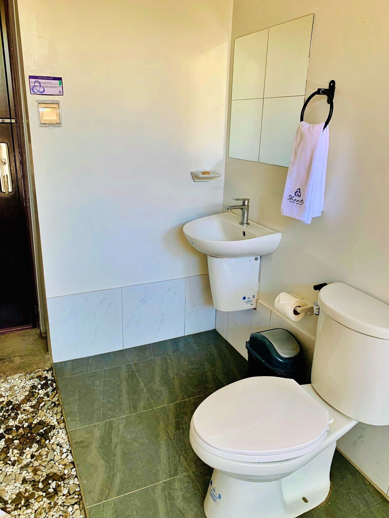 A bathroom with a toilet, sink, and mirror. There is a towel hanging on the wall and a trash bin next to the toilet.