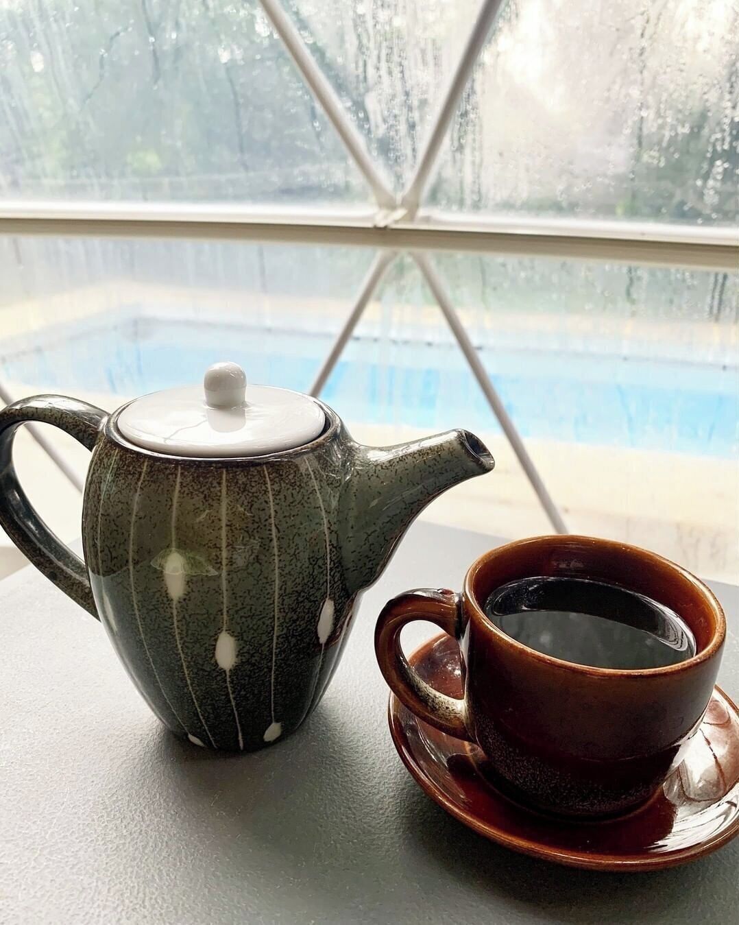 A teapot and cup on a table by a window with a view of a pool.