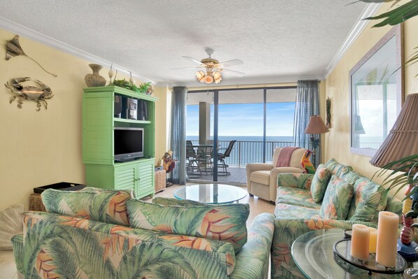 A living room with a green cabinet, a sofa, a glass coffee table, and a view of the ocean through large windows. - 🌊White Caps 1502 | Gulf Front | Awesome Amenities🌊 (Orange Beach)