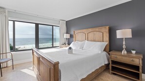 Une chambre avec une grande fenĂȘtre donnant sur la plage, un lit en bois, deux tables de chevet avec des lampes et un fauteuil.