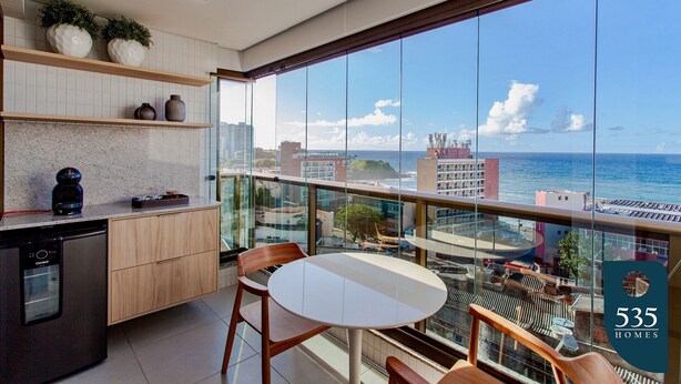 A modern kitchen with a dishwasher, wooden cabinets, and a dining area with a table and chairs. A large window offers a view of the ocean and cityscape. - Barra with Sea View - Cozy and Well Decorated (Salvador)