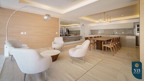 Un saló modern amb un sofà, una butaca i una taula de centre. Hi ha una zona de menjador amb una taula i cadires. El paviment és de fusta i el sostre té il·luminació empotrada.