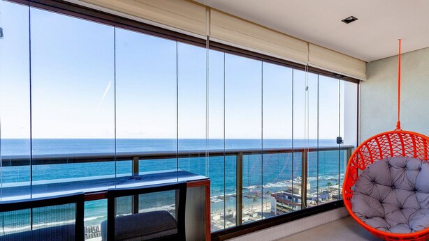 A balcony with a view of the ocean, a hanging chair, and a small table. - 2 Bedrooms with Home Office Sea View in Barra (Salvador)