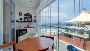 A balcony with a wooden table and chairs, a kitchen area with a refrigerator, and a view of the beach and a building with a conical roof.