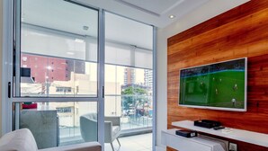 A modern living room with a large window, a flat-screen TV mounted on a wooden wall, a sofa, and a small table with a remote control.