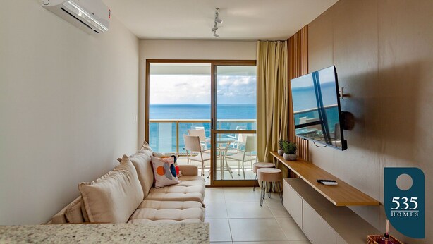 Een moderne woonkamer met een bank, een aan de muur bevestigde televisie, een balkon met uitzicht op zee en een plafondventilator.