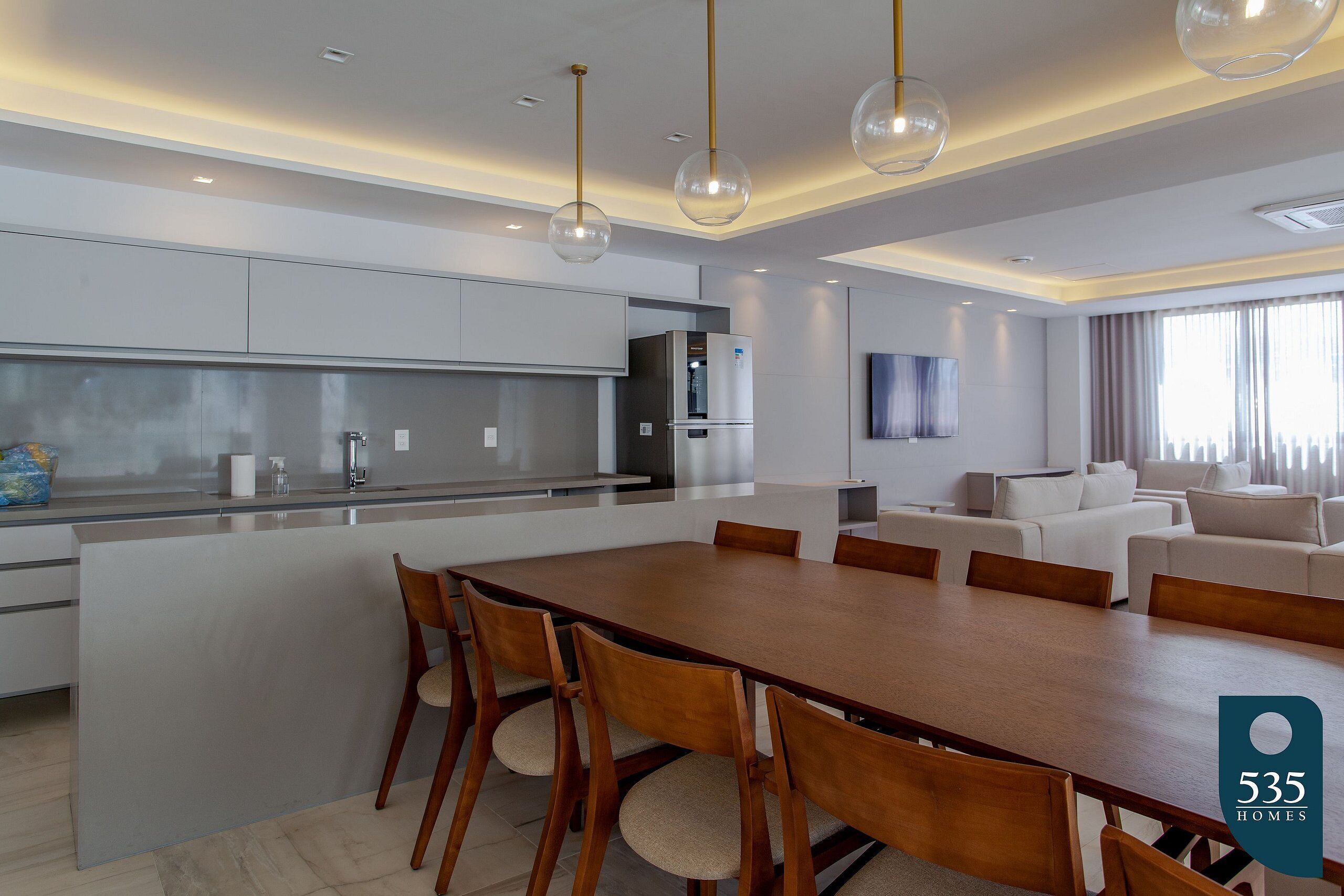 A modern kitchen with a central island, dining table, and chairs. There is a living area with a sofa and a TV.