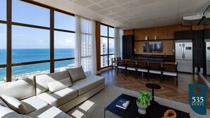 Una sala de estar moderna con un ventanal grande con vistas al océano, un sofá, un comedor y una cocina con electrodomésticos de acero inoxidable.