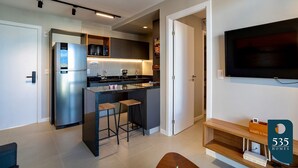 Una cocina moderna con refrigerador empotrado, microondas y una isla con taburetes de bar. Un televisor de pantalla plana está montado en la pared, y hay un banco de madera con almacenamiento debajo.