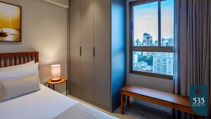 Habitación de hotel con cama, mesita de noche con lámpara, armario, ventana con vista a la ciudad y un banco.