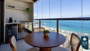 A modern kitchen with a view of the ocean, a dining area with a round table, and a small refrigerator. - Experience the Charm of Barra Near the Lighthouse (Salvador)