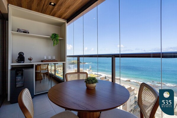 A modern kitchen with a view of the ocean, a dining area with a round table, and a small refrigerator. - Experience the Charm of Barra Near the Lighthouse (Salvador)