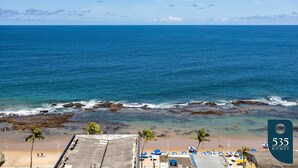 A beach with clear blue water, a rocky shoreline, and a few people. There are umbrellas and lounge chairs on the beach. - Experience the Charm of Barra Near the Lighthouse (Salvador)