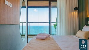 Habitación de hotel con un ventanal que ofrece vista al mar, una cama y una toalla rosa, además de un cartel con el nombre del hotel.