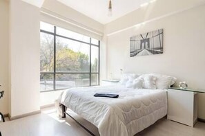 Un dormitorio moderno con un ventanal grande, una cama con ropa de cama blanca, una mesita de noche y una obra de arte colgada en la pared.