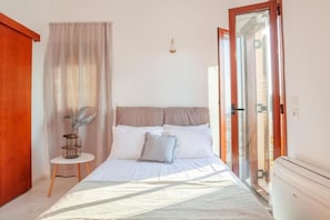 Un dormitorio con cama, una mesita con un florero, una ventana con cortinas y una puerta.