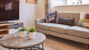 A living room with a sofa, two decorative pillows, a coffee table with plants, a TV, and a window with curtains.