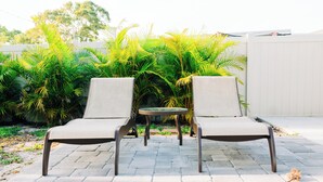 Two lounge chairs and a small table on a paved patio with a white fence and green plants in the background. - A Modern Beach Concept - Re:Treat 408 (Bradenton)